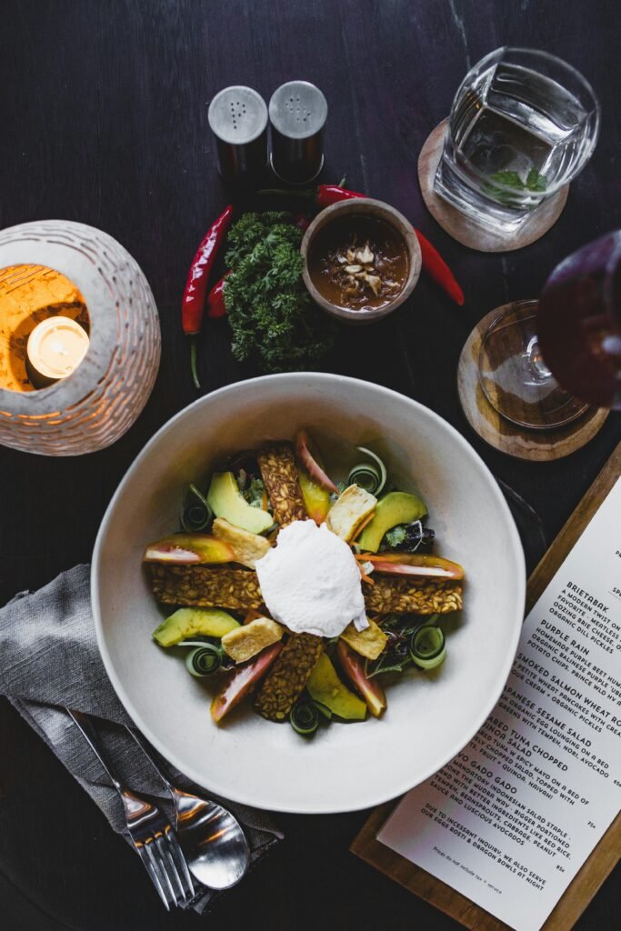 Top view of ceramic bowl with appetizing salad with avocado slices and crisps with fried egg white in center near glass of water and candle with menu in restaurant