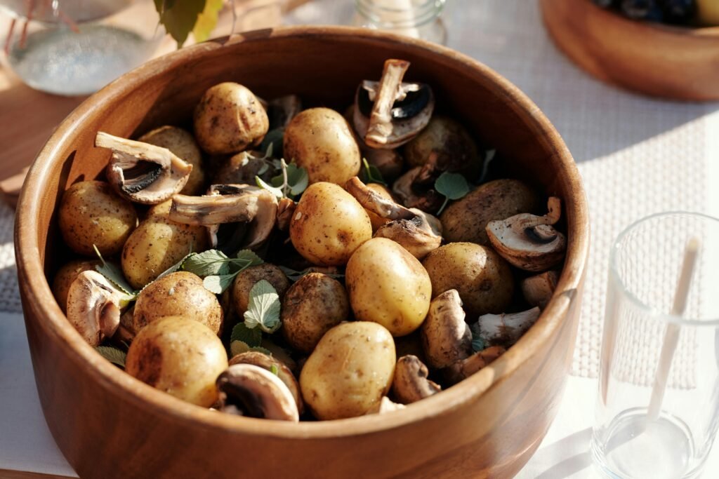 A wooden bowl filled with fresh potatoes, mushrooms, and herbs in natural lighting.